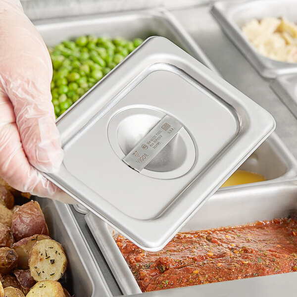 A hand holding a Vigor stainless steel hotel pan cover over food.