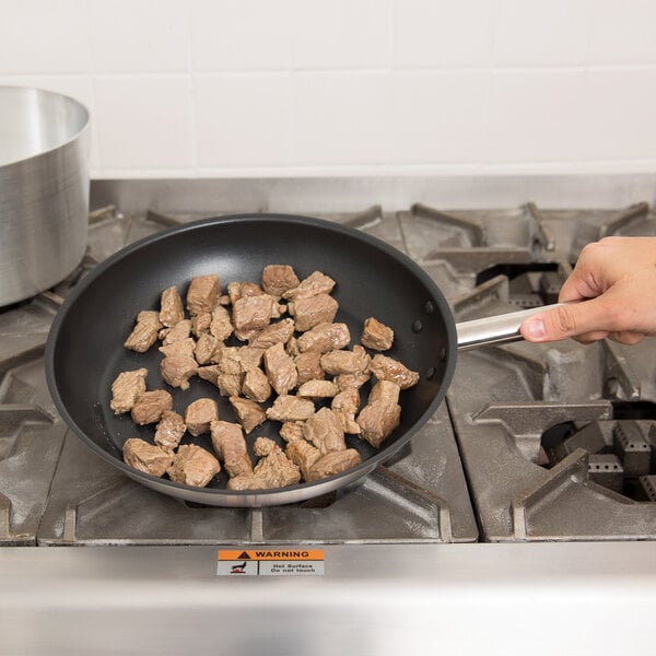 A hand holding a Vollrath stainless steel non-stick fry pan with meat cooking in it.