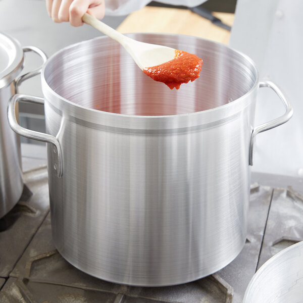 A person stirring red sauce in a Vollrath stock pot with a wooden spoon.