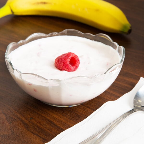 An Arcoroc glass bowl filled with yogurt with a raspberry on top, next to a spoon and a banana.