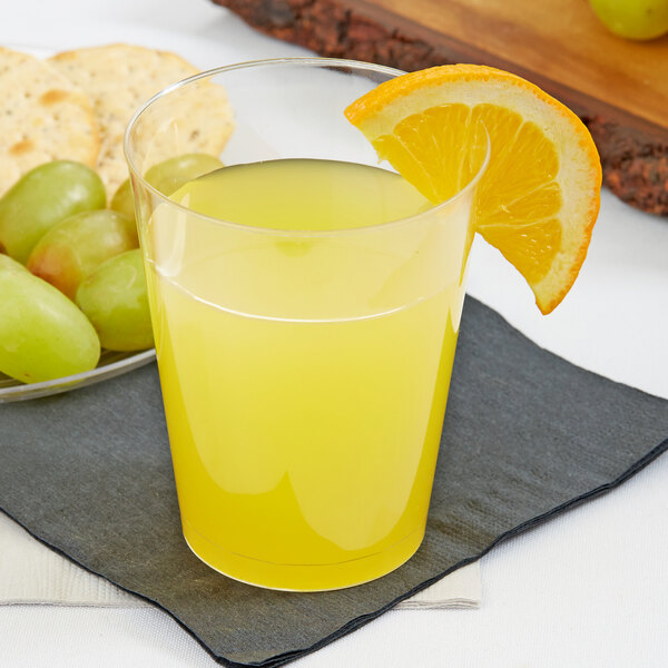 A Fineline tall clear hard plastic tumbler filled with orange juice with a slice of orange on the rim, surrounded by grapes and crackers.