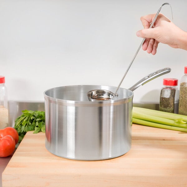 A hand using a metal spoon to stir soup in a Vollrath aluminum sauce pan with a TriVent handle.