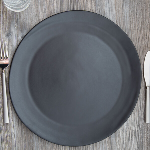 A black 10 Strawberry Street Ripple charger plate with silverware on a wooden table.