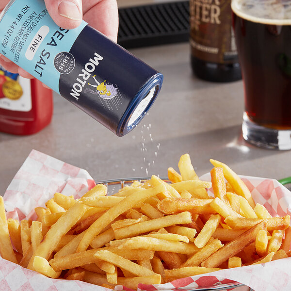 A hand holding a Morton Fine Sea Salt can over a basket of french fries.