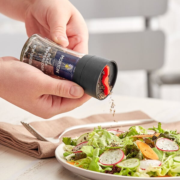 A hand using a Morton Black Peppercorn Mill to pour pepper onto a salad with vegetables.