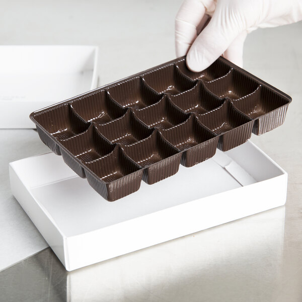 A gloved hand holding a brown plastic candy tray filled with chocolates.