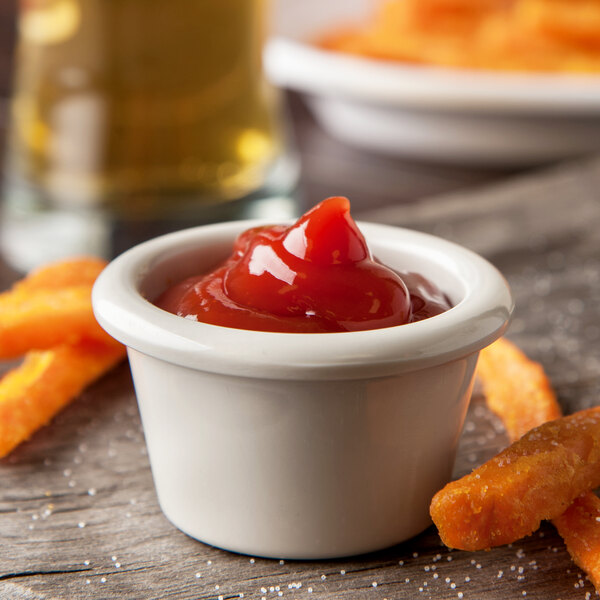 A Carlisle ivory bone ramekin filled with ketchup on a table with french fries.