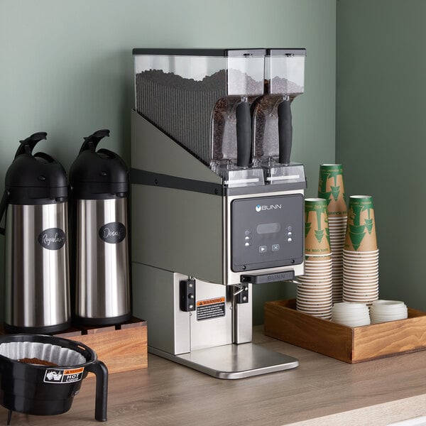 A Bunn BrewWISE multi hopper coffee grinder on a counter with coffee cups and beans in it.