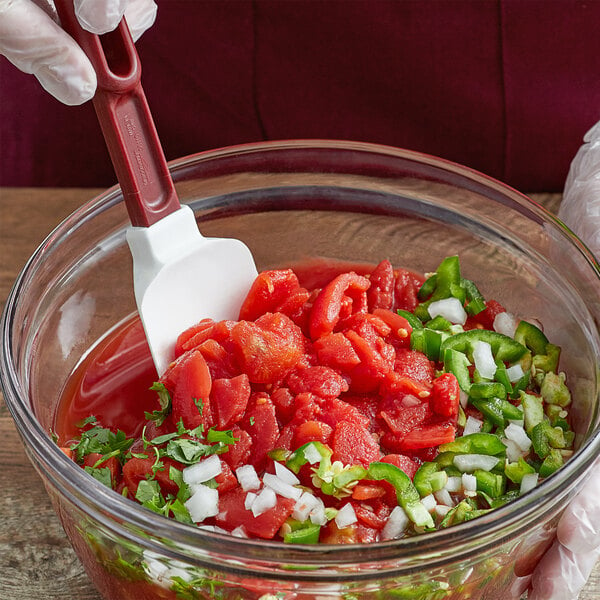 A gloved hand mixes chopped vegetables in a bowl of Furmano's diced tomatoes with a red spatula.