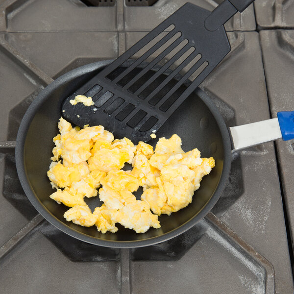 A Vollrath aluminum non-stick fry pan with scrambled eggs and a spatula on a stove.