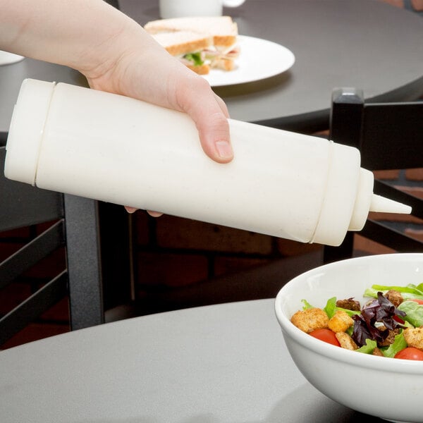 A hand using a Tablecraft dualway squeeze bottle to drizzle sauce over a bowl of salad with tomatoes and croutons.