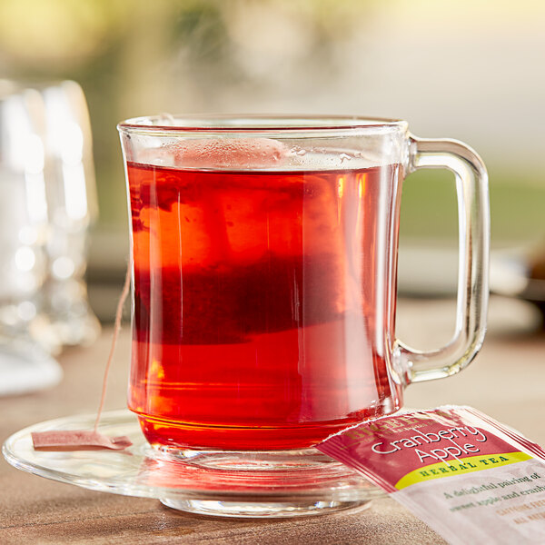 A glass mug with red Bigelow Cranberry Apple herbal tea on a saucer with a tea bag.