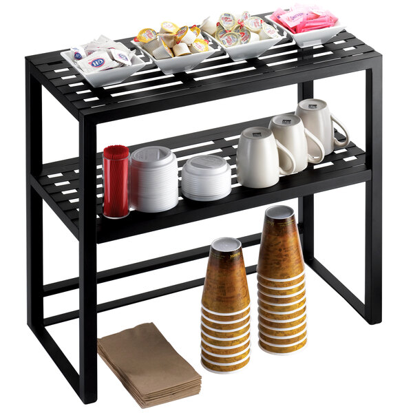 A Cal-Mil black metal two tier display shelf holding white mugs, bowls, and plates.