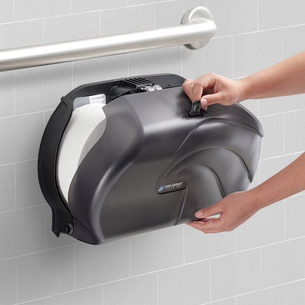 A hand holding a San Jamar black plastic double roll toilet paper dispenser.