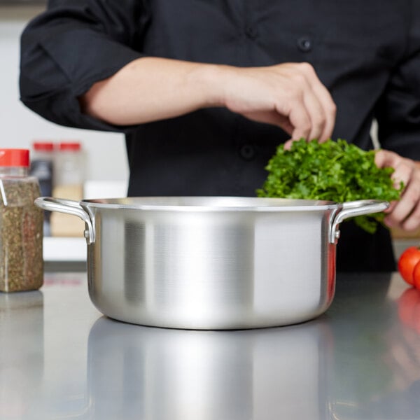 A chef in a black shirt uses a Vollrath stainless steel sauce pot to cook green vegetables.