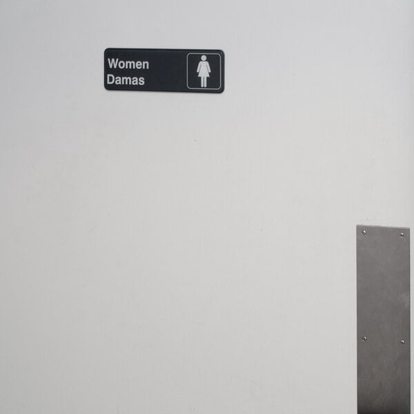 A white sign with black text and a woman figure reading "Women's Restroom" on a grey metal wall.