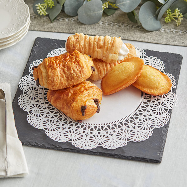 A stack of white plates topped with a plate of pastries with a Hoffmaster Cambridge lace doily.