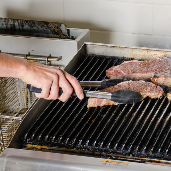 A hand using Vollrath Jacob's Pride tongs with a piece of meat on a grill.