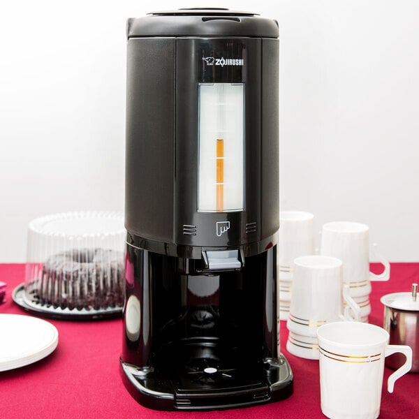 A 2.5 liter gravity container on a table with a coffee maker and white mugs.