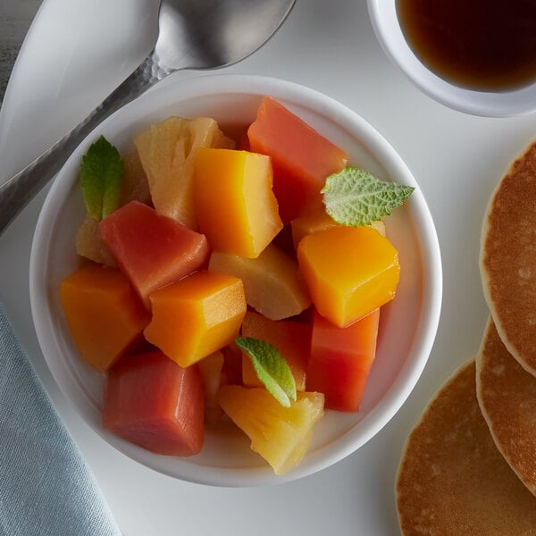 A bowl of tropical fruit salad with a spoon on a table with pancakes and a cup of coffee.
