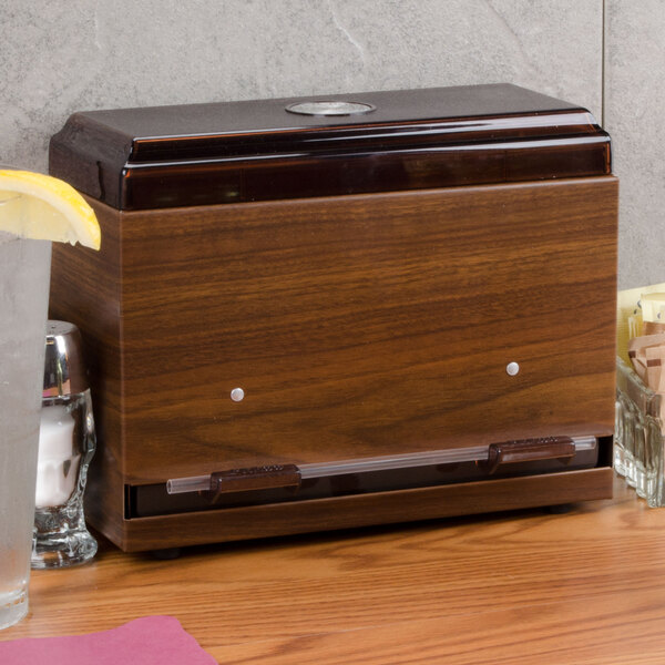 A dark walnut woodgrain Vollrath Straw Boss dispenser on a counter with a glass of water next to it.