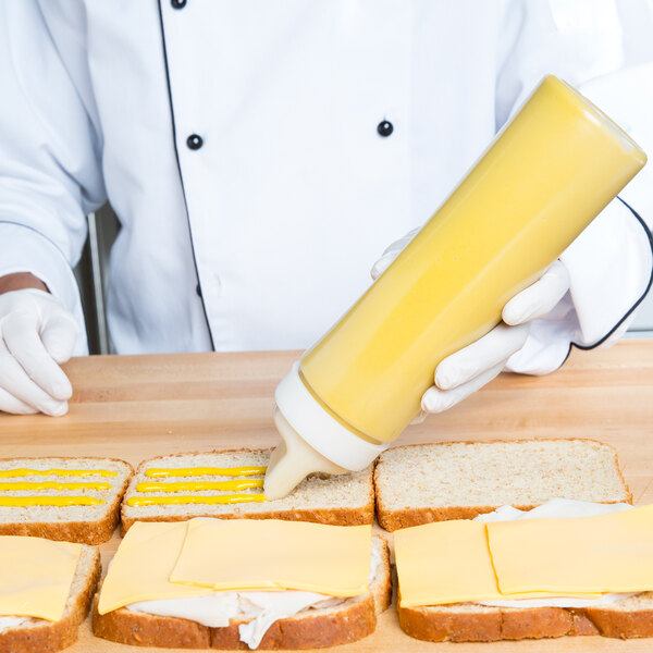 A person using a Tablecraft wide mouth squeeze bottle to pour mustard on a sandwich.