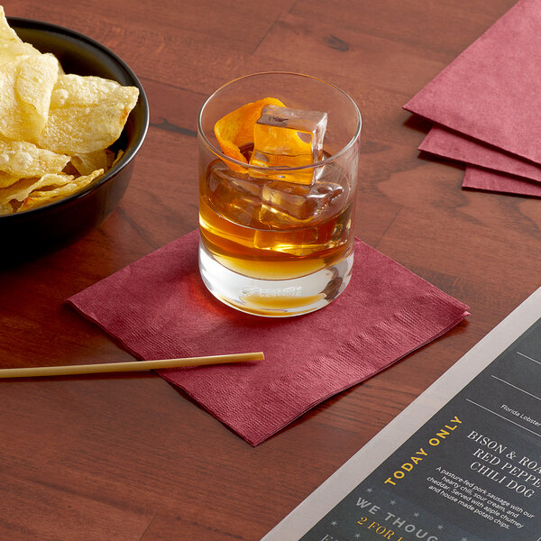A bowl of chips and a drink on a table with a burgundy cocktail napkin.