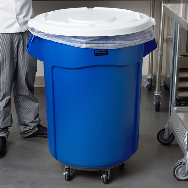 A man standing next to a blue Rubbermaid trash can with a white lid.