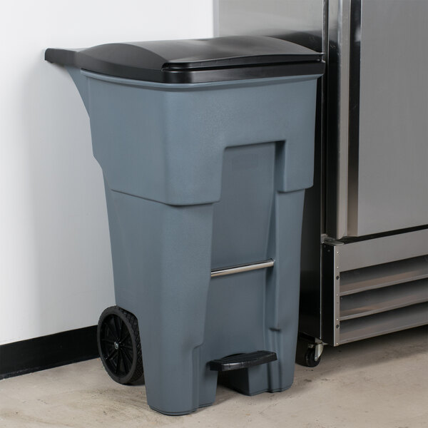 A Rubbermaid grey rectangular trash can with black lid next to a refrigerator.