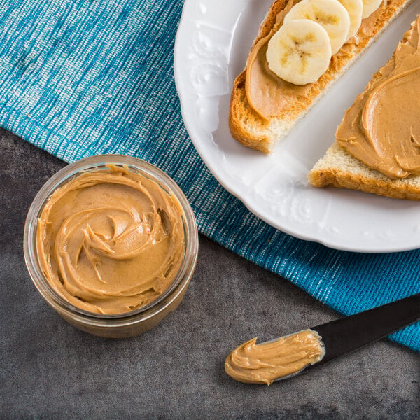 A white plate with toast topped with peanut butter and bananas, a sliced banana, and a jar of Bulk Creamy Peanut Butter.