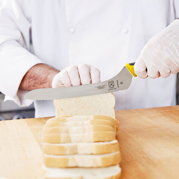 A hand using a Mercer Culinary Millennia Colors yellow bread knife to slice bread on a cutting board.