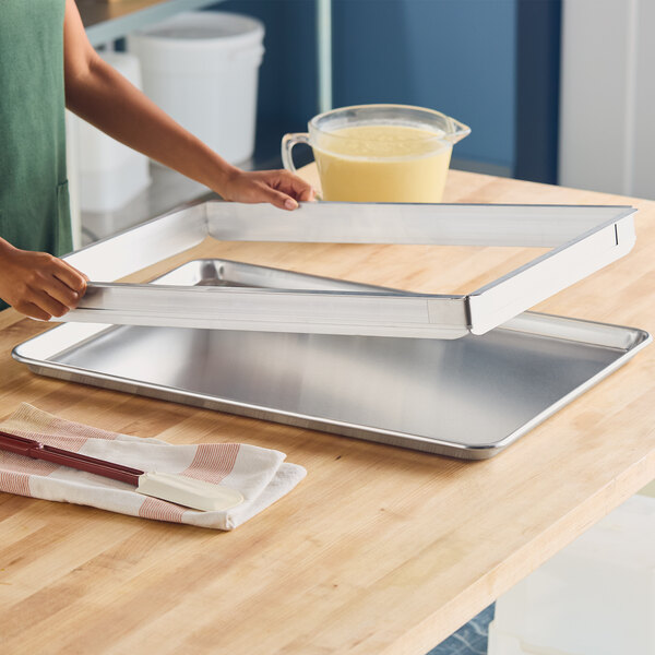 A person is placing a rectangular metal frame onto a large metal baking sheet on a wooden countertop, with a pitcher of batter and utensils nearby.