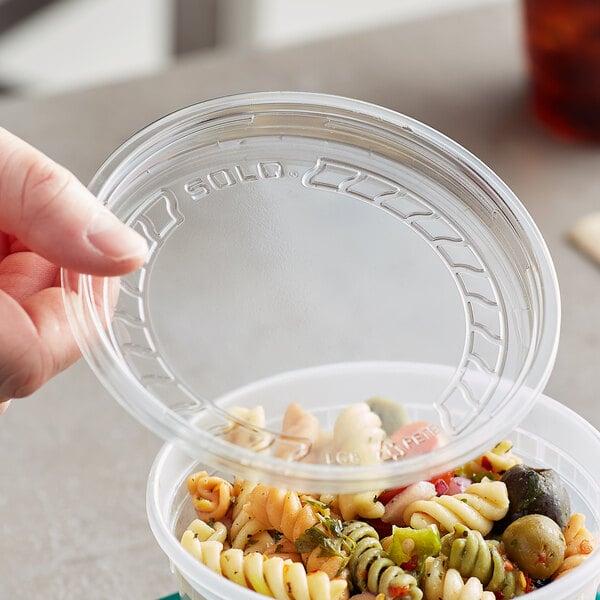 A hand holding a Bare by Solo clear plastic deli container with pasta and olives.