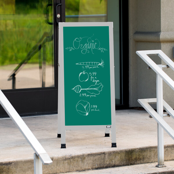 An Aarco green chalk A-Frame sign board with white chalk drawings on it on a sidewalk.