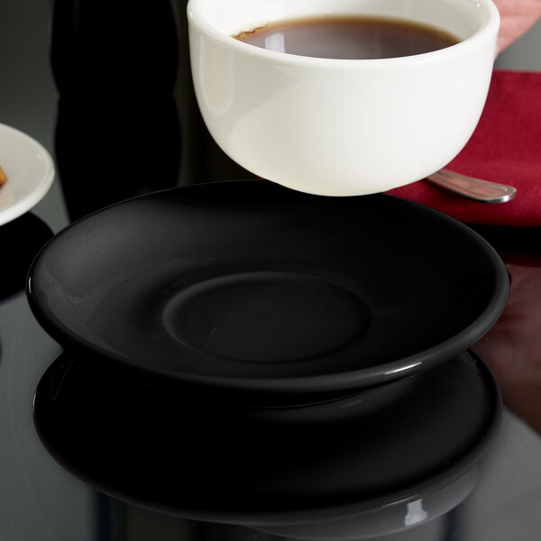 A Tuxton black china saucer with a cup of coffee on a black plate.