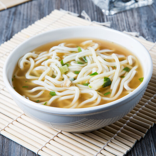 A white Thunder Group melamine bowl filled with noodles in soup with chopsticks and a spoon.