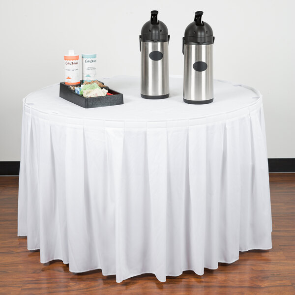 A table with a white Snap Drape Wyndham table skirt and two metal containers on it.