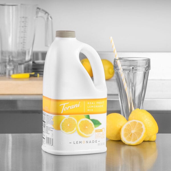 A white jug of Torani Lemonade Smoothie Mix on a counter next to a glass of lemons.