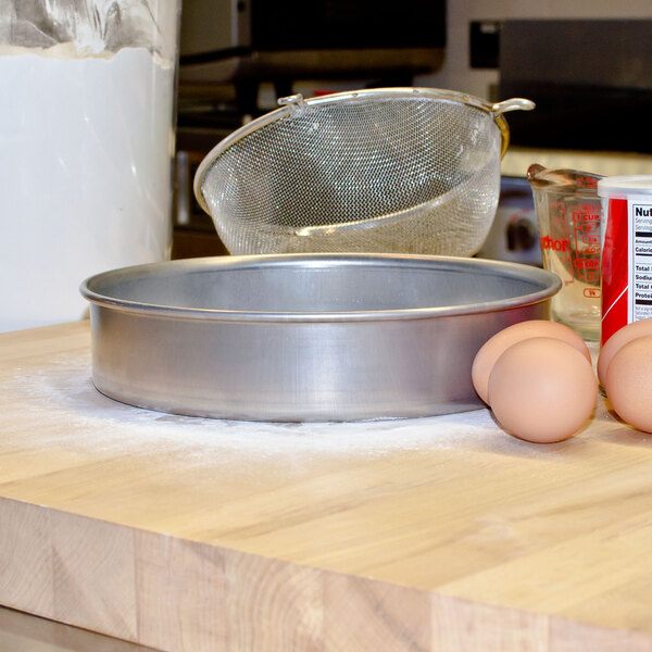 A Vollrath round aluminum cake pan with eggs and flour on a wooden surface.