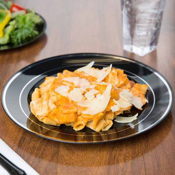 A Fineline black plastic plate with silver bands holding pasta with cheese and salad next to a glass of water.