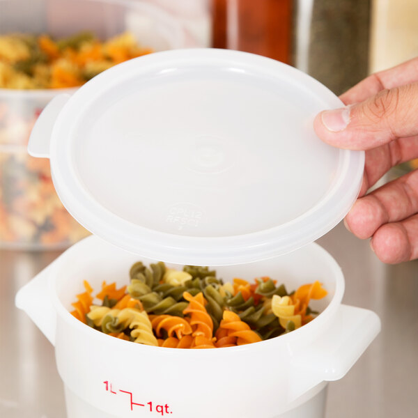 A hand holding a Cambro white plastic lid over a white container of multicolored pasta.