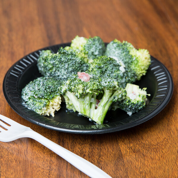A Dart black foam plate with a piece of broccoli on it, next to a fork on a wood table.