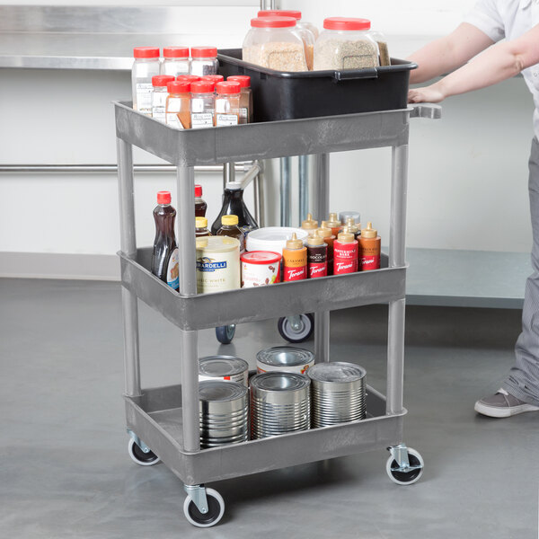 A person holding a Luxor gray plastic utility cart with three tiers of food items on it.