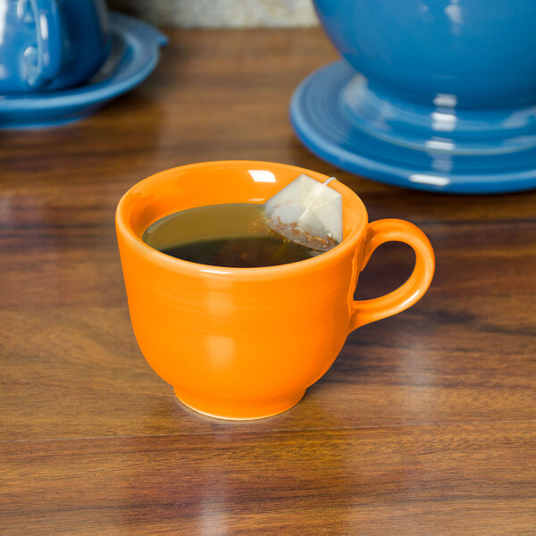 A Fiesta Tangerine china cup filled with tea and a tea bag on a table.