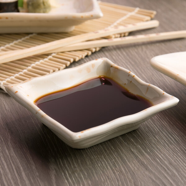 A table with a white square Melamine sauce dish filled with soy sauce next to chopsticks.