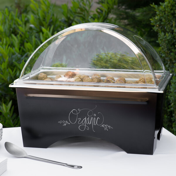 A black Sterno chalkboard chafer with a clear lid on a table with food inside.