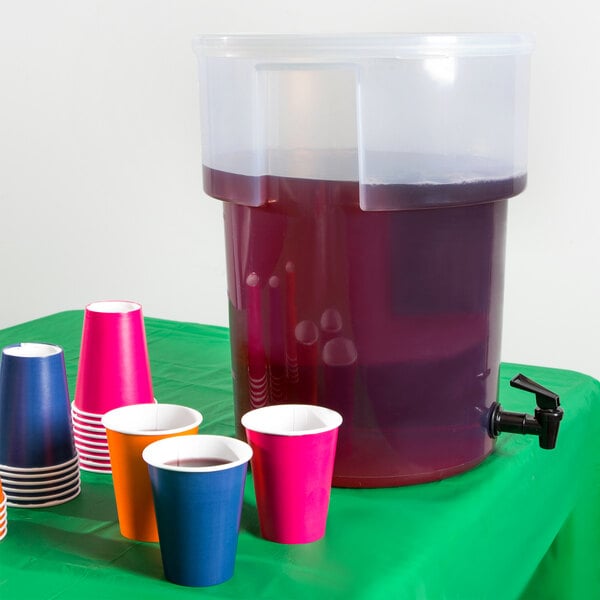 A Carlisle round plastic beverage dispenser with a purple liquid inside and cups on a table.
