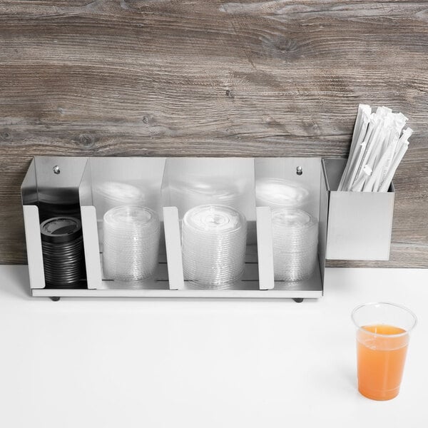 A Vollrath stainless steel wall mount/countertop lid holder with straw holder holding plastic cups and straws on a countertop.