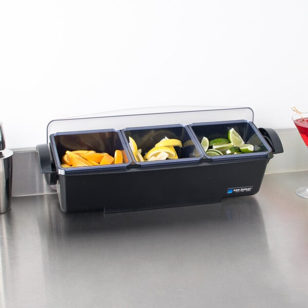 A San Jamar 3-compartment condiment bar on a counter with containers of fruit in it.