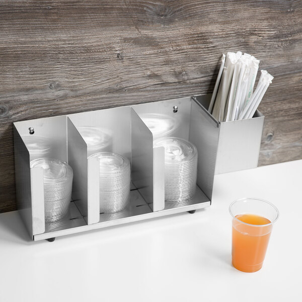 A Vollrath stainless steel lid holder with straw holder on a counter with plastic cups and a glass of juice.